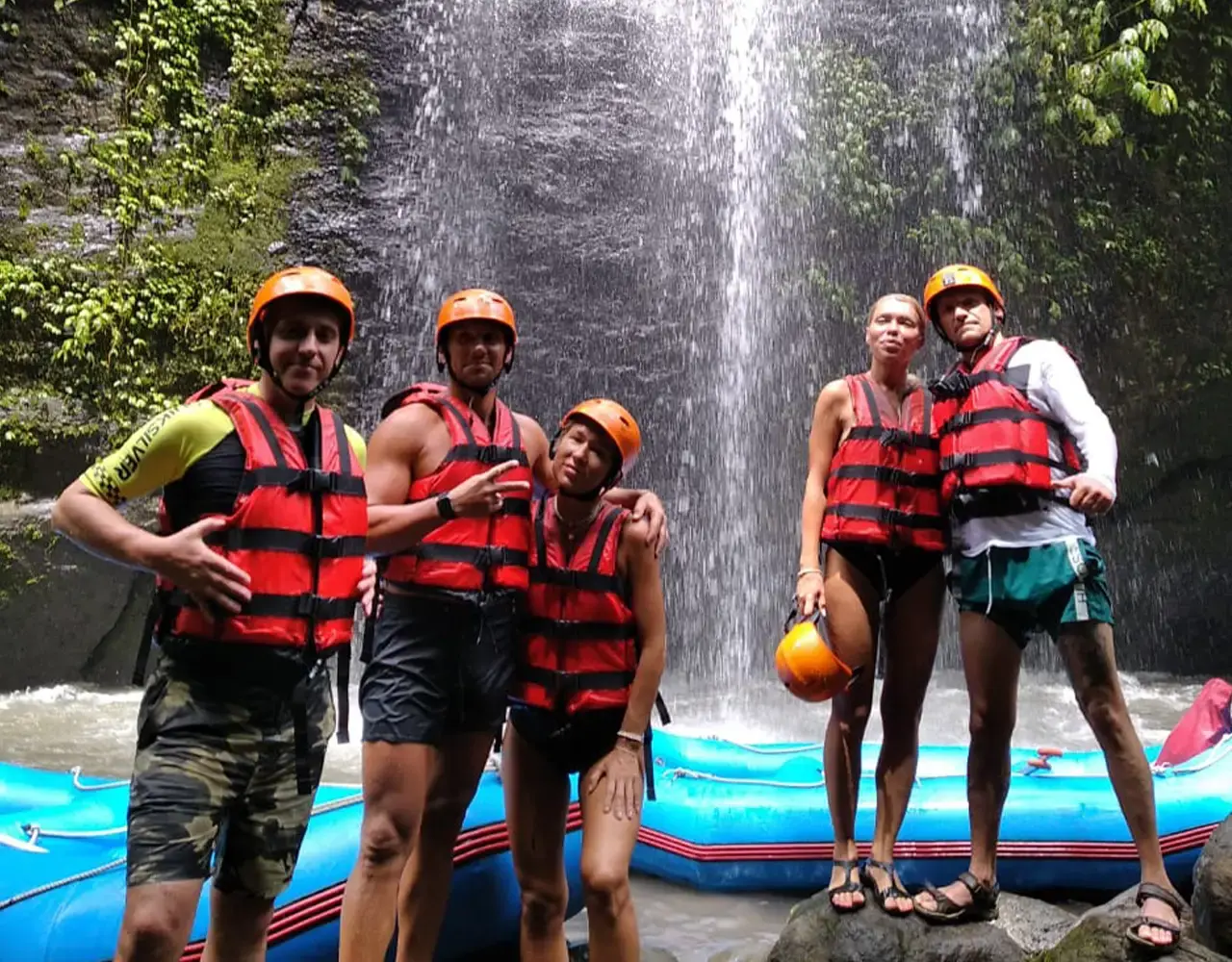 Five rafters in life jackets and helmets standing in front of a waterfall beside their raft