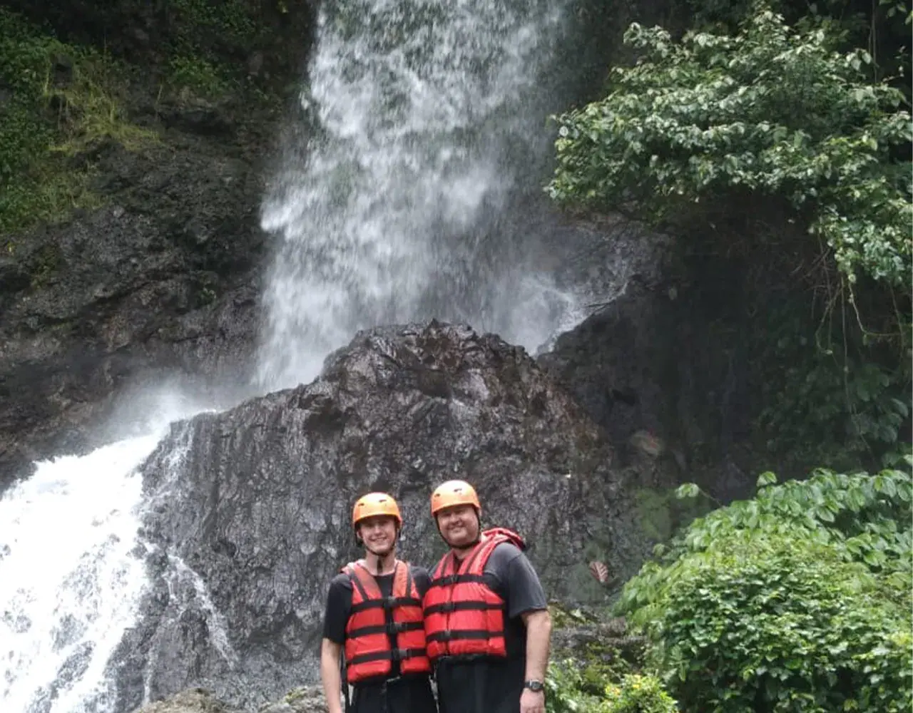 Two rafters in helmets and life jackets standing below a waterfall on the Telaga Waja route